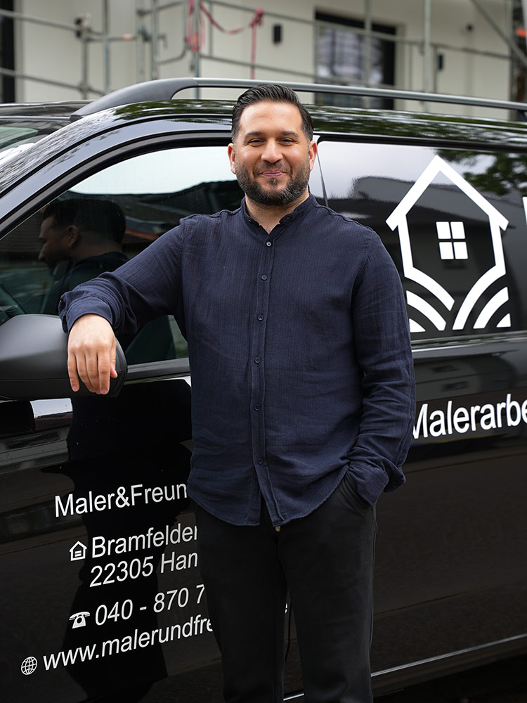 Mann in schwarzem Hemd lächelt, lehnt an schwarzem Firmenauto, Text und Logos sichtbar.