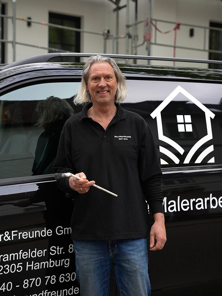 Ein lächelnder Mann mit Pinsel, schwarzes Hemd, vor Firmenfahrzeug mit Hauslogo.