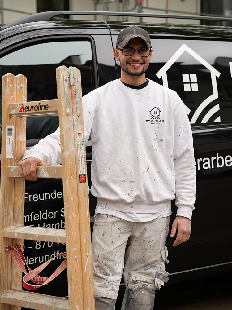 Ein Mann mit Brille und Mütze lächelt, steht neben einer Holzleiter vor einem schwarzen Fahrzeug.
