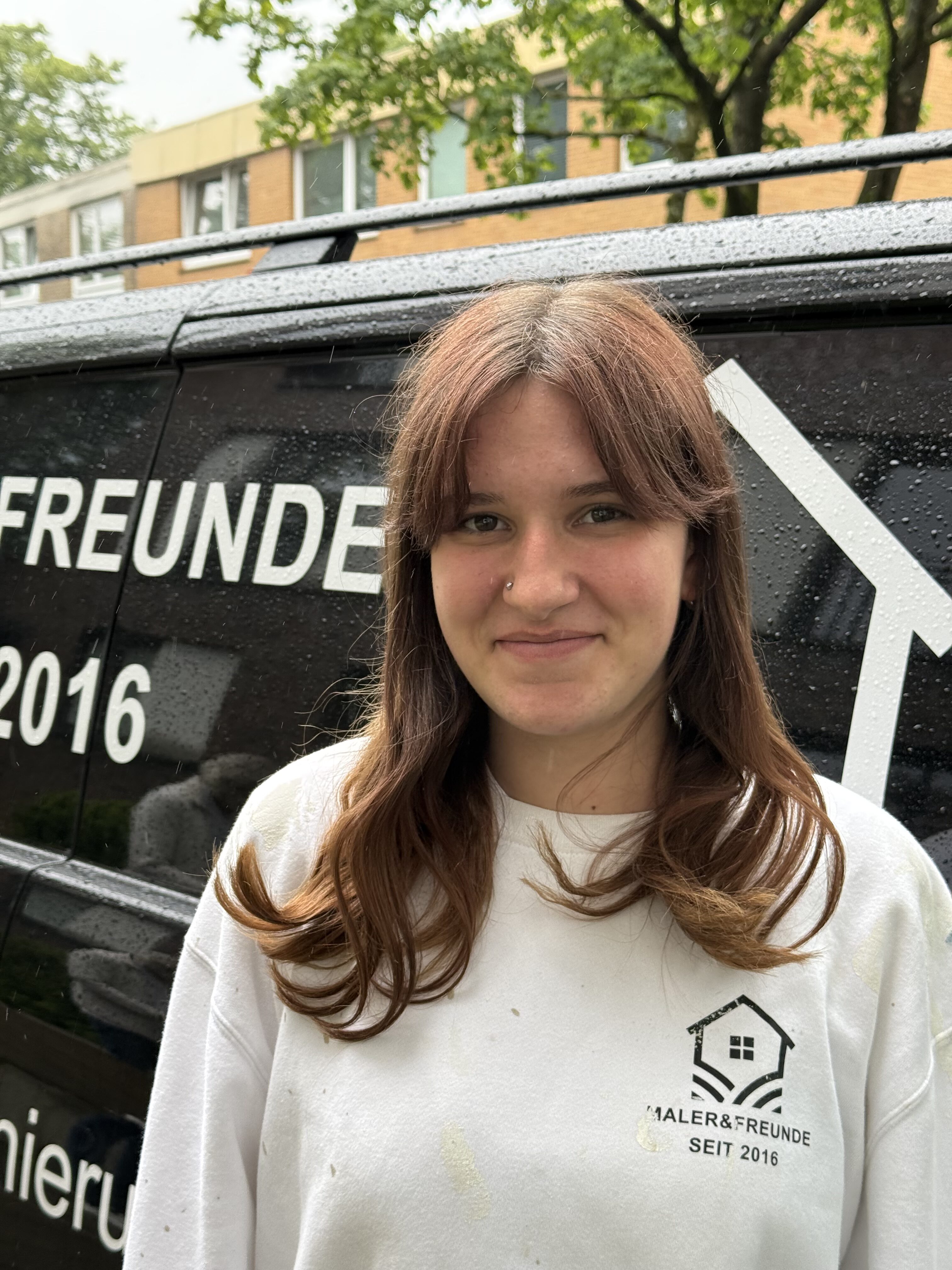 Eine Frau mit langen braunen Haaren lächelt leicht vor einem schwarzen Fahrzeug.